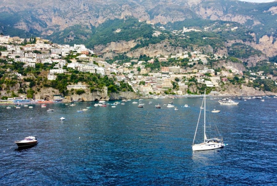 Aerial view of a yacht sailing in the turquoise waters of the Amalfi Coast, surrounded by lush green landscapes and clear skies, captured by a drone.